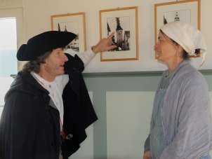 Turmwächterführung in Neunkirch Eine Person mit schwarzem Umhang und schwarzem Hut zeigt auf ein Bild mit einem Turm der brennt darauf. Eine zweite Person mit einem Hellen Kleid und weissem Kopftuch hört gespannt zu.