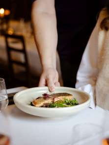 Ein Teller mit einem hübsch angerichteten Fischgericht wird auf einen weiss gedeckten Tisch gestellt. Auf dem Tisch stehen auch mehrere Weingläser mit Rosé.