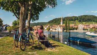 Velofahren Stein am Rhein Zwei Freudinnen sitzen auf einer Holzbank am Rhein mit Blick auf Stein am Rhein. Hinter ihnen stehen zwei E-Bikes.