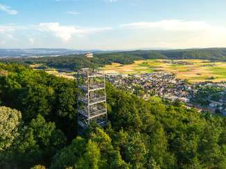 Ein eckiger Metallturm ragt aus dem Wald auf einem Hügel. Unten sieht man auf das Dorf Beringen.