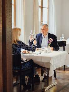 Ein Paar sitzt an einem weiss gedeckten Tisch und stosst mit Wein an. Auf dem Tisch steht ein schön angerichtetes Essen.