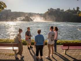 Rheinfallführung Eine Gruppe von Menschen steht am Rheinfallbecken. Im Hintergrund ist der Rheinfall zu sehen. Alle hören gespannt einer Person zu.