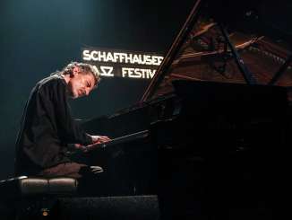 Schaffhauser Jazzfestival Ein Mann sitz an einem Flügel und spielt auf einer Bühne. Im Hintergrund sieht man ein Leuchtschild mir der Schrift "Schaffhauser Jazz Festiaval".