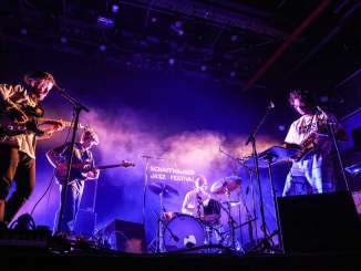 Schaffhauser Jazzfestival Auf einer Bühne stehen ein Schlagzeuger, zwei E-Gitarristen und ein Saxophonist in violettem Scheinwerferlicht.