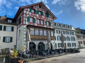Rheingerbe Stein am Rhein Aussenansicht eines Riegelhauses. Vor dem Haus stehen viele Tische und Stühle sowie drei grosse Sonnenschirme. Das haus hat grüne Fensterläden und einen langen Holzerker.