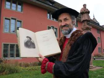 Ein Mann mit schwarzem Umhang hält ein Buch hoch. Auf der linken Buchseite ist ein altes gemaltes Portrait zu sehen. Im Hintergrund ist eine rote Statue und eine rote Hausfassade zu sehen.