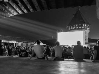 Viele Personen sitzen am Boden oder auf Bänken und schauen einen Film auf einer grossen Leinwand, die vor dem Turm des Munots steht.