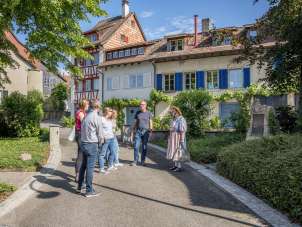 Hallauer Gässliwanderung Eine Frau in einer Tracht steht vor einer Gruppe und erzählt Geschichten. Die Gruppe steht in einer Gasse mit Häusern mit bunten Fassaden.