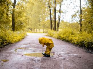 Ein Kind trägt eine gelbe Regenjacke und Regenhose und schwarze Gummistiefel. Es spielt in eine Regenpfütze auf einem Waldweg.