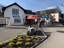 Eine Tankstelle mit einem Blumenbeet im Vordergrund. Die Zapfsäulen sind blau. Es steht in grosser Schrift, dass die Tankstelle bedient ist. Daneben gibt es ein Kiosk.