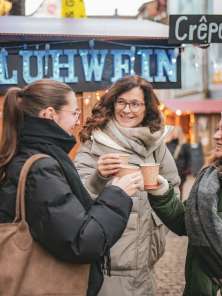 Drei Freudinnen tragen dicke Winterkleidung, stehen vor einem Glühweinstand und stossen mit einem Becher Glühwein an.