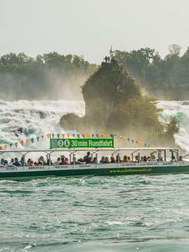 Ein grünes Schiff gefüllt mit Personen. Auf dem Schild auf dem Dach des Schiffes steht: 30 min Rundfahrt. Ebenfalls ist das Schiff mit www.schiffmaendli.ch angeschrieben. Im Hintergrund ist ein Felsen zu sehen.