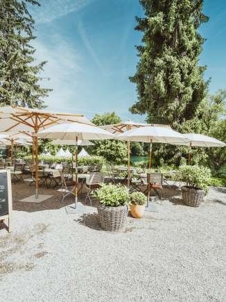 Holzstühle sowie Tische sind gedeckt und weisse Sonnenschirme spenden Schatten. Die Restaurantsterrasse liegt direkt am Rhein mit viel Grün umgeben.