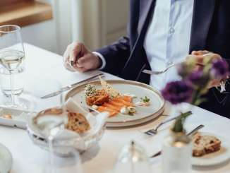 Auf einem weiss gedeckten Tisch steht eine Vorspeise mit Lachs sowie ein Glas Weisswein und Brot.