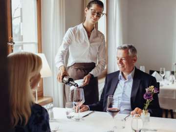 Zwei Personen sitzen an einem weiss gedeckten Tisch. Eine Servicekraft  schenkt ein Glas Rotwein ein.