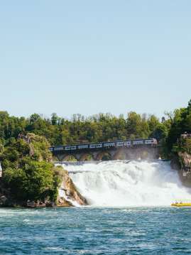 Ein Zug fährt über die Eisenbahnbrücke hinter dem Rheinfall in Richtung Schloss Laufen.