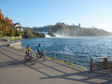 Zwei Velofahrende am Ufer eines Flusses. Im Hintergrund ist ein Wasserfall zu sehen. Am anderen Ufer befindet sich ein Schloss.