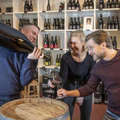 Drei Personen stehen um ein Weinfass. eine Person schenkt mit einer riesigen Weinfalsche Rotwein ein.