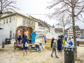 Auf einem grossen Platz steht eine mobile Sauna. Davor stehen Personen im Bademantel.