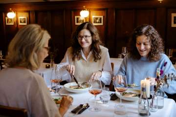 Drei Freundinnen sitzen an einem weiss gedeckten Tisch. Darauf stehen Weingläser mit Rosé und ein Fischmenü.