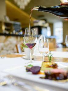 Es wird Rotwein in ein Glas eingeschenkt. Auf dem Tisch steht ein Hauptgang mit einem Stück Fleisch und Beilagen.