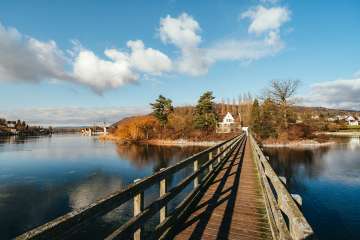 Eine Holzbrücke auf eine Insel mit einem Haus darauf. Im Wasser spiegelt sich der Himmel.