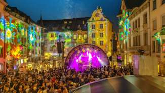 Ein Platz zwischen Häusern. Die Fassaden sind farbig beleuchtet. In der Mitte ist eine Bühne mit Musiker-/innen. Der Platz ist gefüllt mit Menschen.