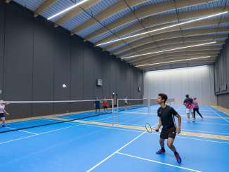 Eine Halle mit vier blauen Badminton-Felder. Darauf spiel Personen Badminton.