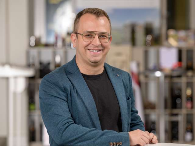Portrait Adrian Provinski. Er trägt einen blauen Sakko und ein schwarzer T-Shirt. Er trägt eine Brille.