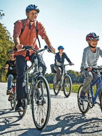 Eine Familie fährt mit dem Velo auf einem Feldweg. Um sie herum hat es Wiesen, Felder und einen Wald.
