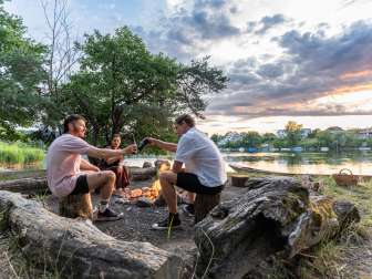 Drei Freunde sitzen bei Abendstimmung am Feuer einer Grillstelle am Rhein und stossen mit einem Bier an.