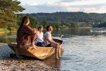 Drei Personen sitzen in einem Weidling am Ufer und geniessen die Abendstimmung am Rhein.