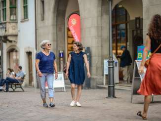 Zwei Freundinnen flanieren auf der Gasse und laufen am Tourist Office vorbei. Es hat ein Schild mit einem blauen "i".