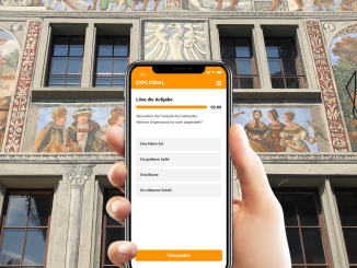 Eine Hand hält ein Hady vor einer mittelalterlichen Fassade. Auf dem Handy muss man ein Rätsel lösen.