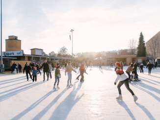 Ein Ausseneisfeld mit Personen, welche Eiskunstlaufen. Am Gebäude dahinter auf dem Schild steht "Eispark KSS".