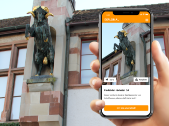 Explorial Schaffhausen Eine Hand hält ein Handy vor ein Gebäude mit einer Statue des Schaffhauser Bocks. Auf dem Handy sieht man das Bild der Statue.