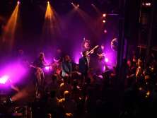 Ein dunkler Raum, eine beleuchtete Bühne mit zwei Sängern und fünf weiteren Musikern. Der restliche Raum ist gefüllt mit Zuschauenden.