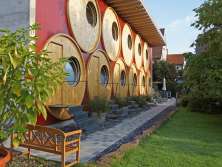 Ein Gebäude mit 11 runden Fässern mit je einem runden Fenster. Davor ein Vorgarten mit Wiese, Bäumen, Steinplatten-Platz und einer Holzbank.