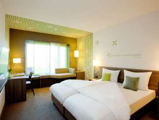 Ein modernes Hotelzimmer mit einem Doppelbett, Fernseher, Arbeitspult und Sitzecke. Das Zimmer ist mit grünen Akzenten dekoriert.