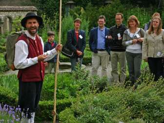 Eine Person mit historischer Bekleidung und einem Holzstock in der Hand sowie eine Gruppe von 7 Personen stehen in einem Kräutergarten. Die Gruppe hört der verkleideten Person gespannt zu.
