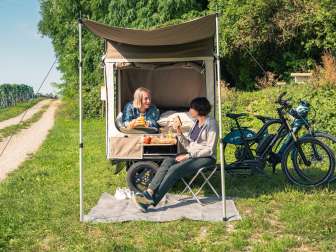 Auf einer Wiese neben Reben ist ein Cargovelo mit aufgeklapptem Zeltaufbau. Eine Peron liegt im Bett und sitz auf einem Campingstuhl davor.