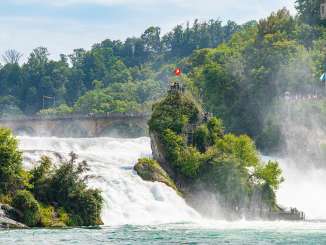 Blick auf einen mächtigen Felsen mitten im Wasserfall. Eine Treppe führt hoch auf eine kleine Plattform, wo Touristen stehen.