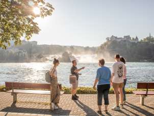 Eine Gruppe von Menschen steht am Rheinfallbecken. Im Hintergrund ist der Rheinfall zu sehen. Alle hören gespannt einer Person zu.