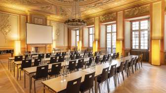 Zunftsaal Sorell Hotel Rüden Ein Saal mit prächtiger Stuckdecke ist für ein Seminar eingerichtet. Vorne steht eine Leinwand und dahinter Tischreihen mit Stühlen.