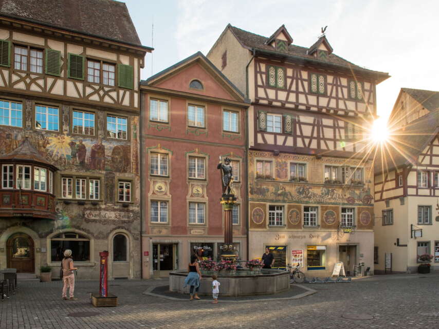 Fassadenmalereien in der Altstadt Stein am Rhein. Die Sonne scheint durch die Häuser. Vor den Häusern steht ein Brunnen.