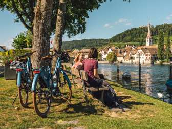 Zwei Freudinnen sitzen auf einer Holzbank am Rhein mit Blick auf Stein am Rhein. Hinter ihnen stehen zwei E-Bikes.