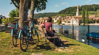 Zwei Freudinnen sitzen auf einer Holzbank am Rhein mit Blick auf Stein am Rhein. Hinter ihnen stehen zwei E-Bikes.