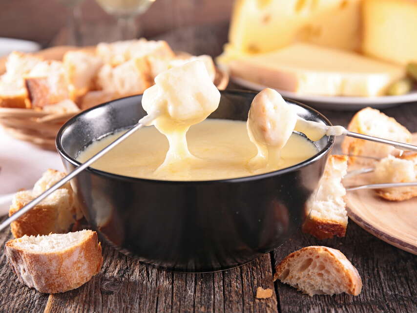 Käsefondue. Zwei Gabel tunken ihr Brot im Käsefondue. Im Hintergrund steht ein Korb mit Brotstücken und ein angeschnittener Käse.