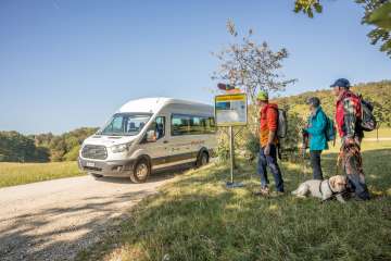 Drei Wanderer und ein Hund warten an einer Bushaltestelle. Ein Kleinbus hält in diesem Moment und hat eine Aufschrift "Clientis Randenbus".