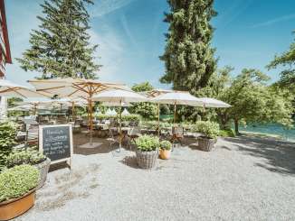 Terrasse Gasthof Schupfen Holzstühle sowie Tische sind gedeckt und weisse Sonnenschirme spenden Schatten. Die Restaurantsterrasse liegt direkt am Rhein mit viel Grün umgeben.